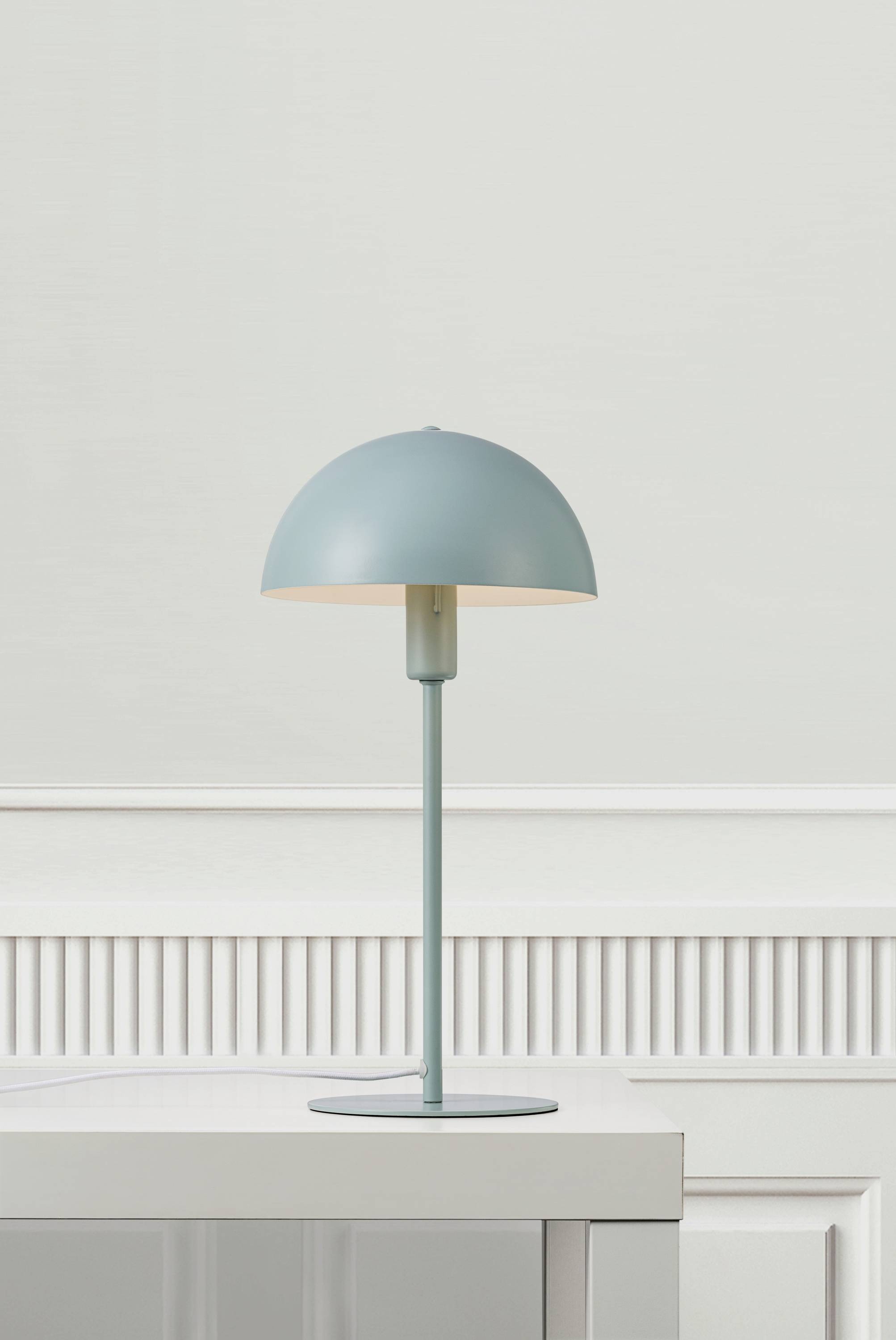 A minimalist table lamp with a blue, semi-spherical lampshade sits on a white table in front of a light-coloured wall.