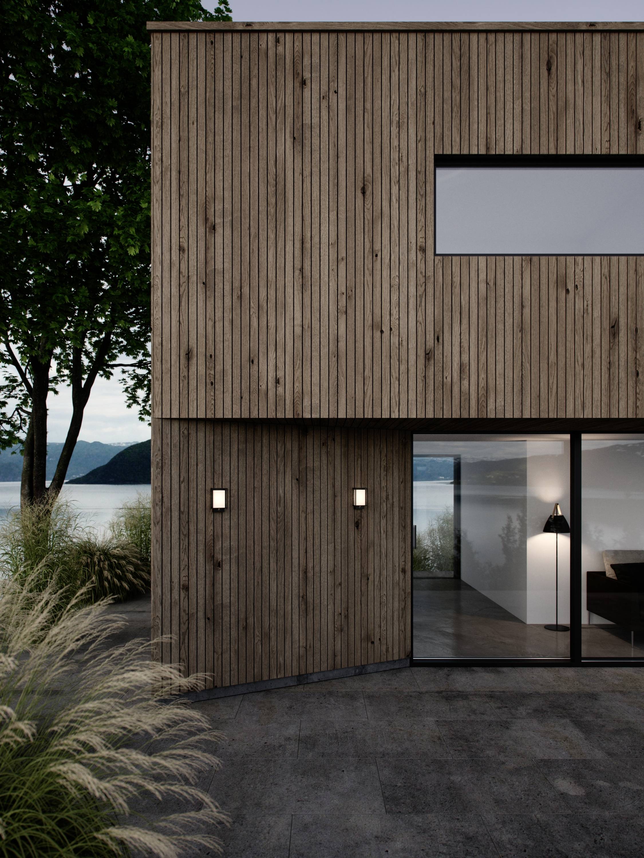 Modern wooden house with large windows, surrounded by plants, overlooking a tranquil lake and mountains in the background.