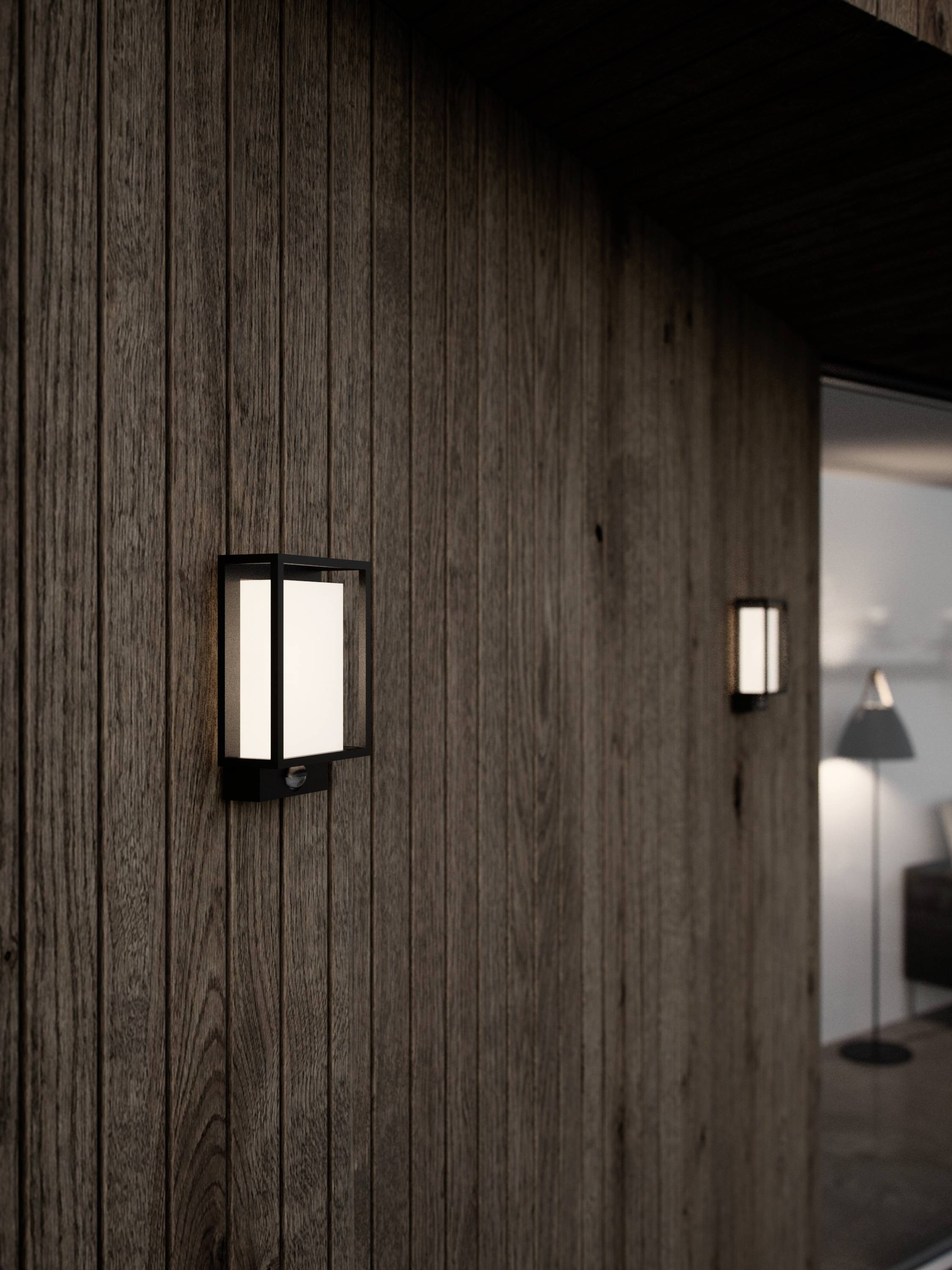Two wall-mounted exterior lights illuminate a wooden cladding. In the background, a living room with a standard lamp is blurrily visible.