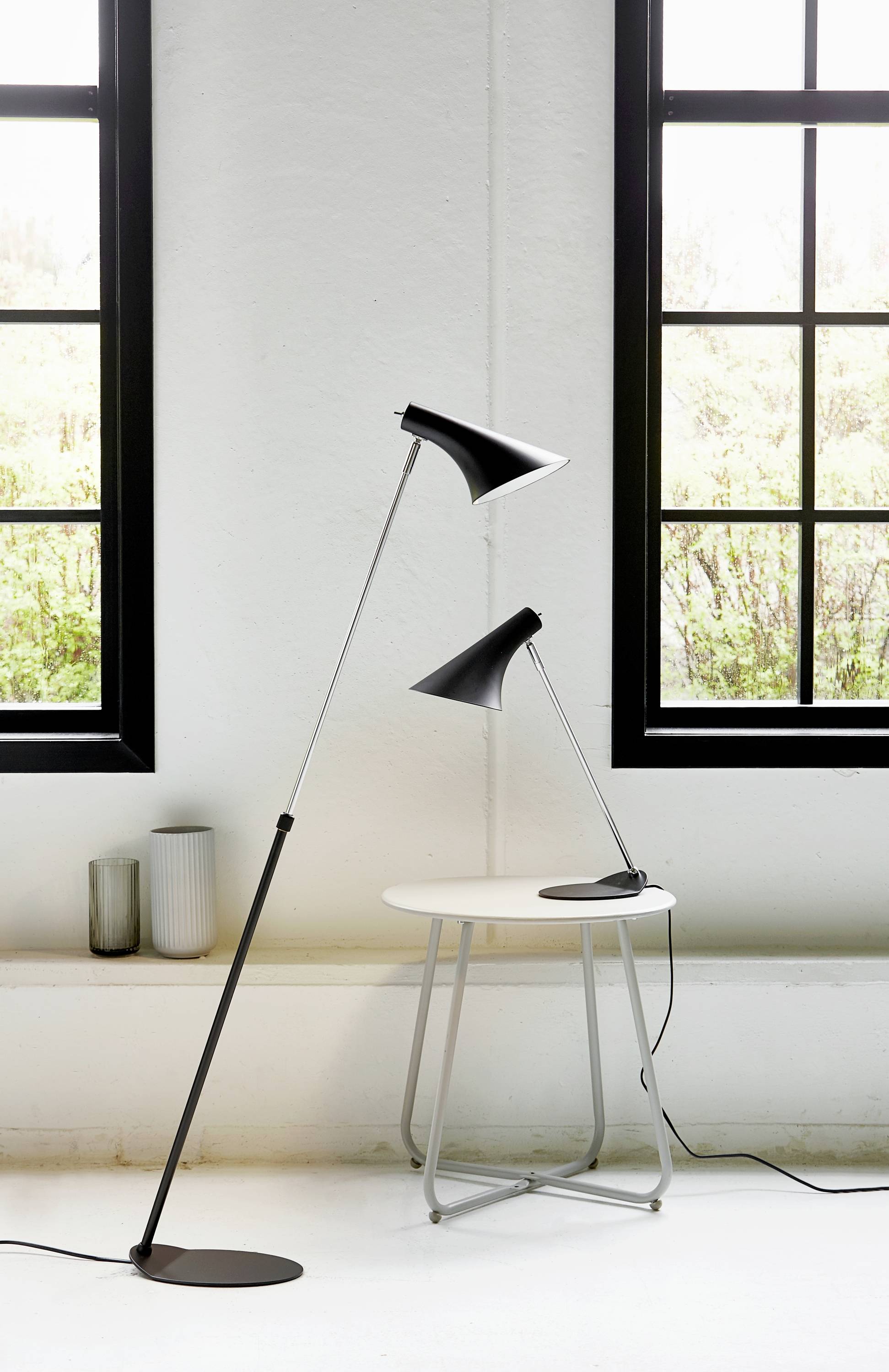 Two black designer lamps are placed on and beside a small white table, in front of a window with a black frame. Minimalist interior.