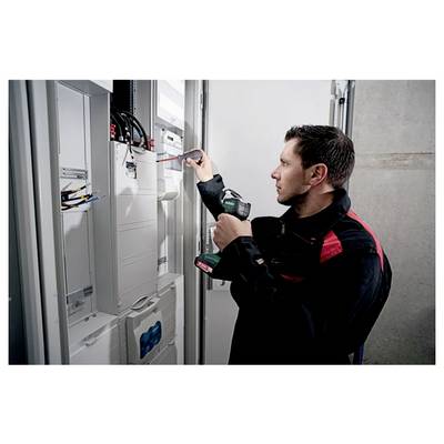 A technician in uniform uses a screwdriver and a drill to work on electrical panels in a control room.