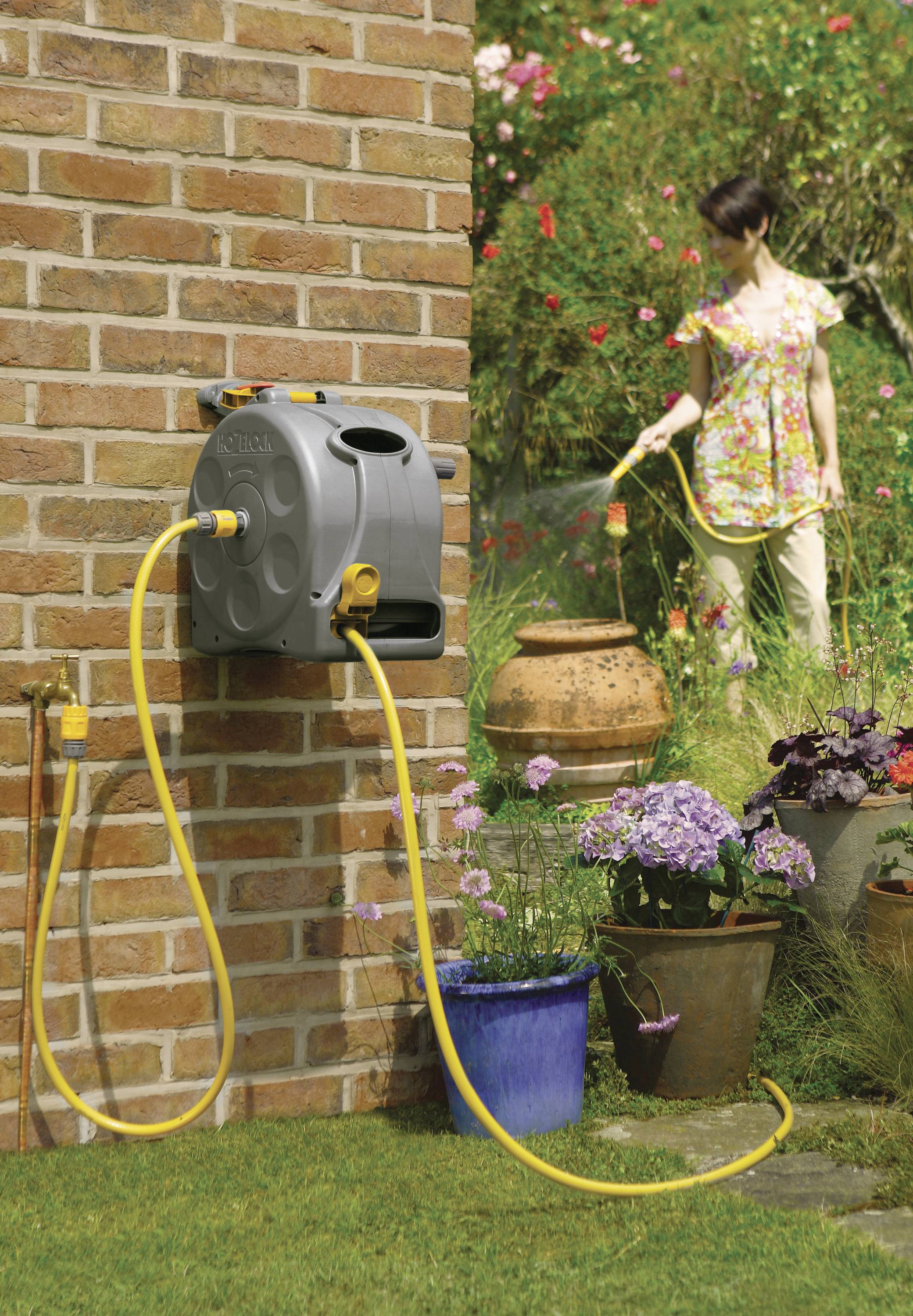 A woman is watering plants in the garden with a hosepipe attached to a wall mount. Flower pots are in the foreground.