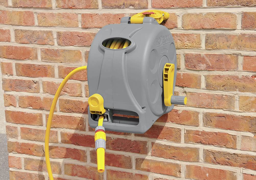 Grey hose reel with yellow garden hose, mounted on a red brick wall. Crank on the side for winding up the hose.