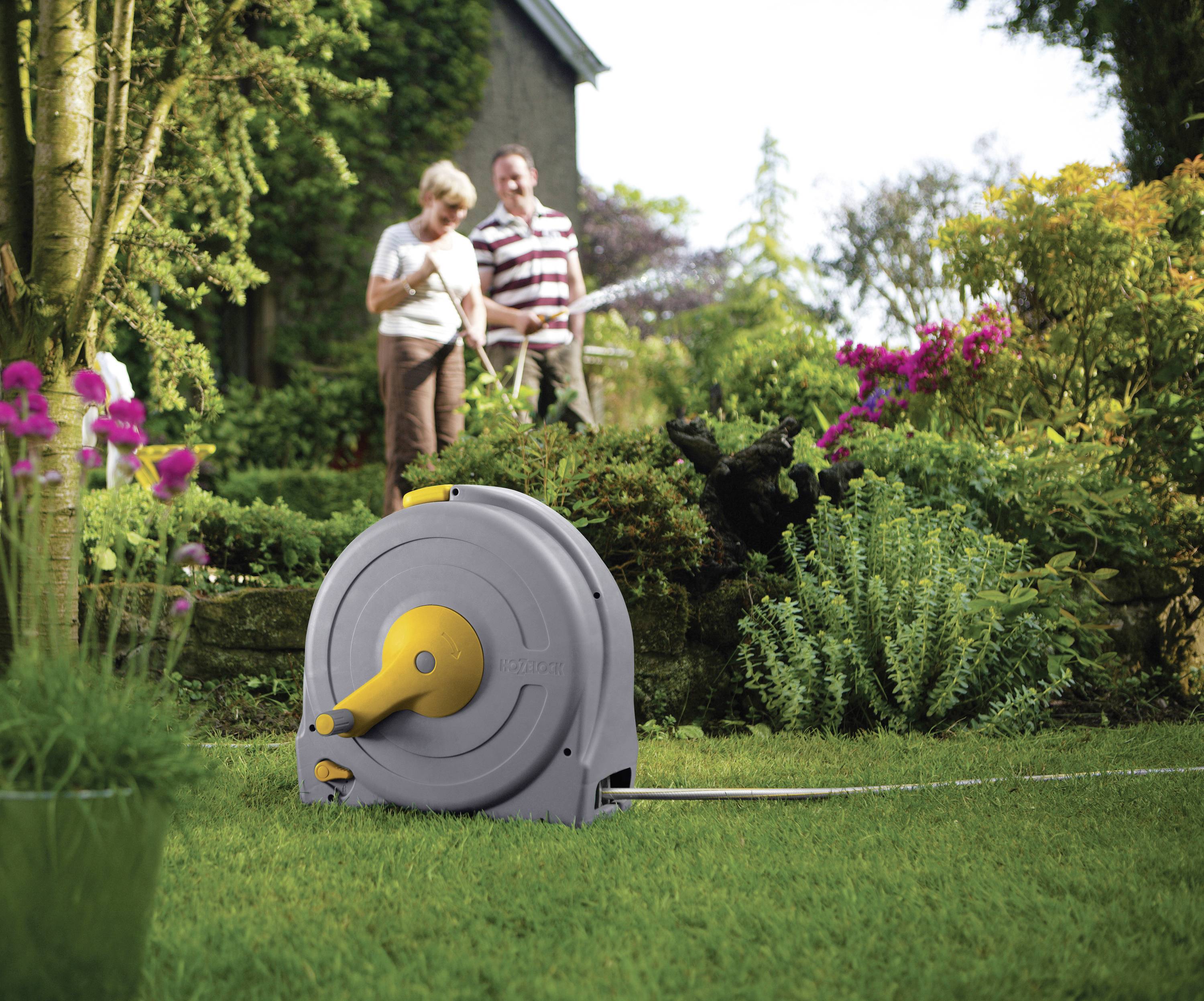 An elderly couple waters the garden with a hosepipe from a ground-standing hose reel. Flowering plants are visible in the background.
