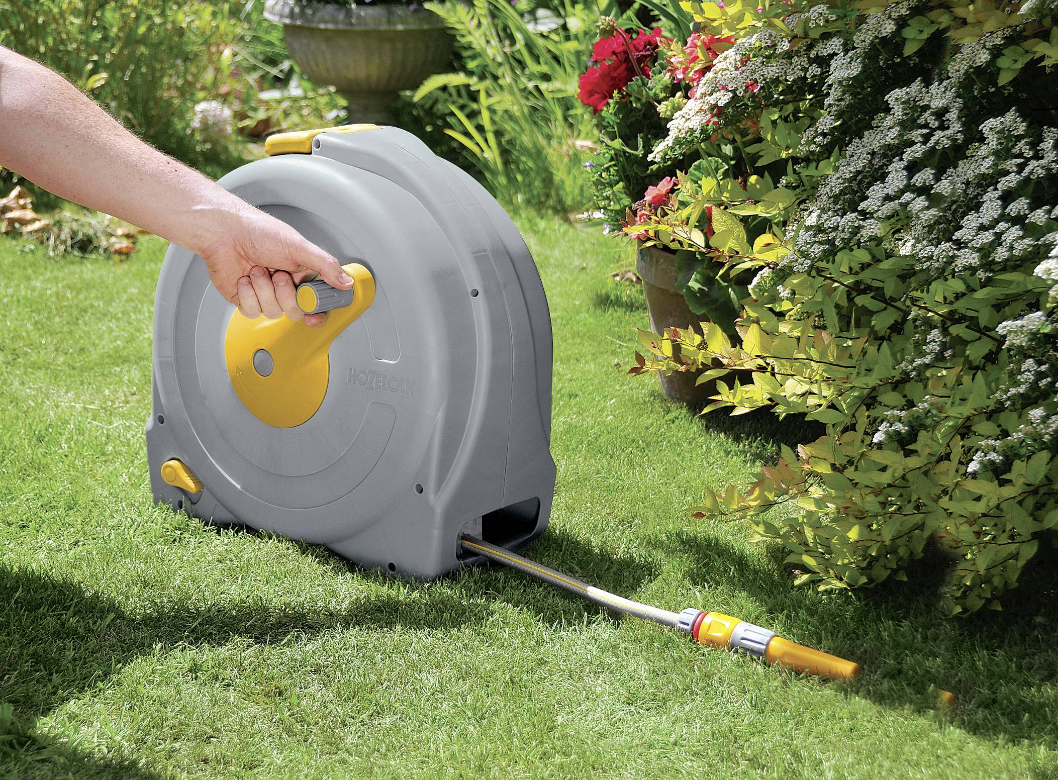 A person is operating a garden hose reel on a green lawn, surrounded by flowers and shrubs.