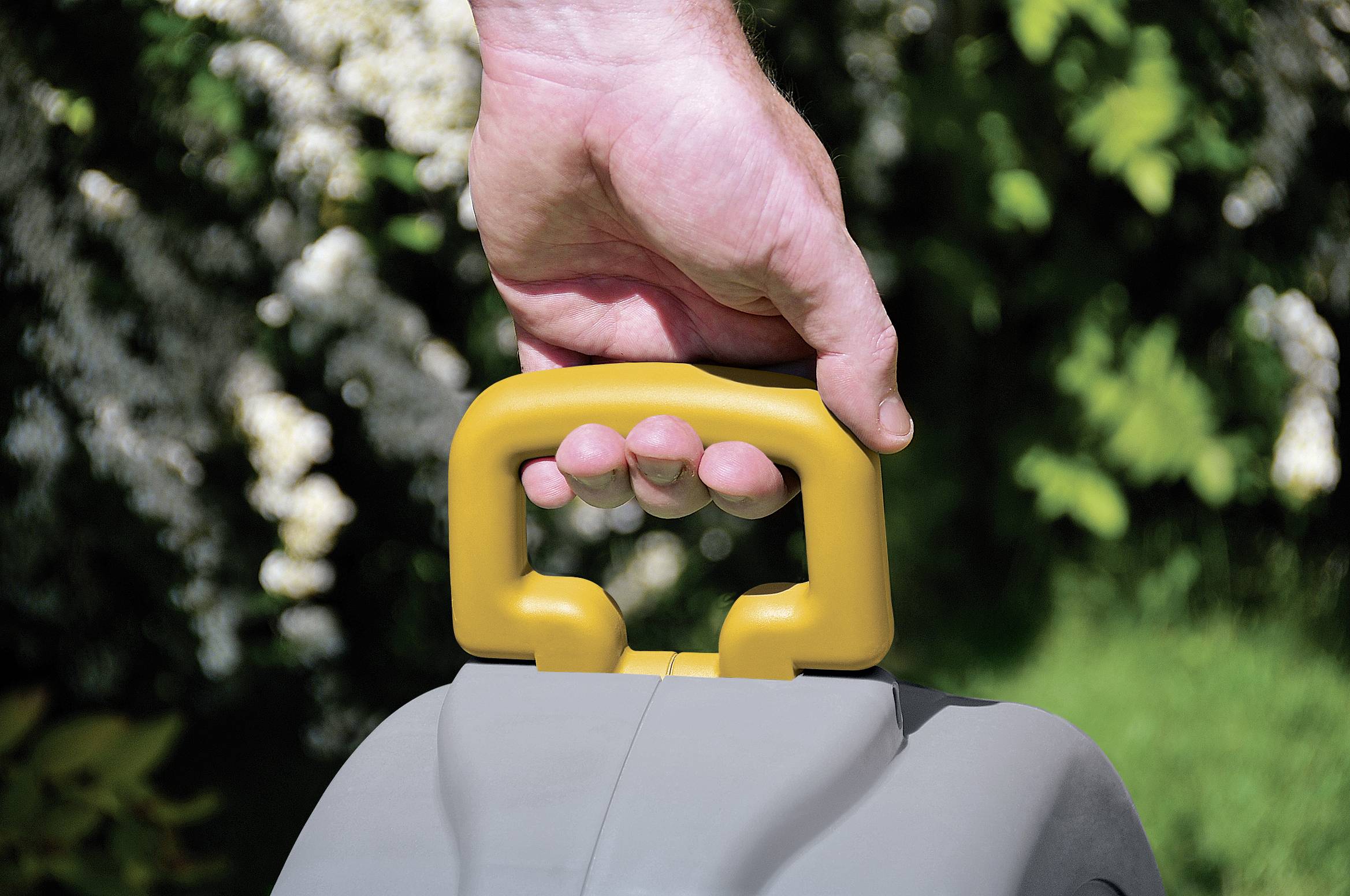 A person is holding the yellow handle of a grey garden tool. The background consists of blurred green and white-flowering plants.