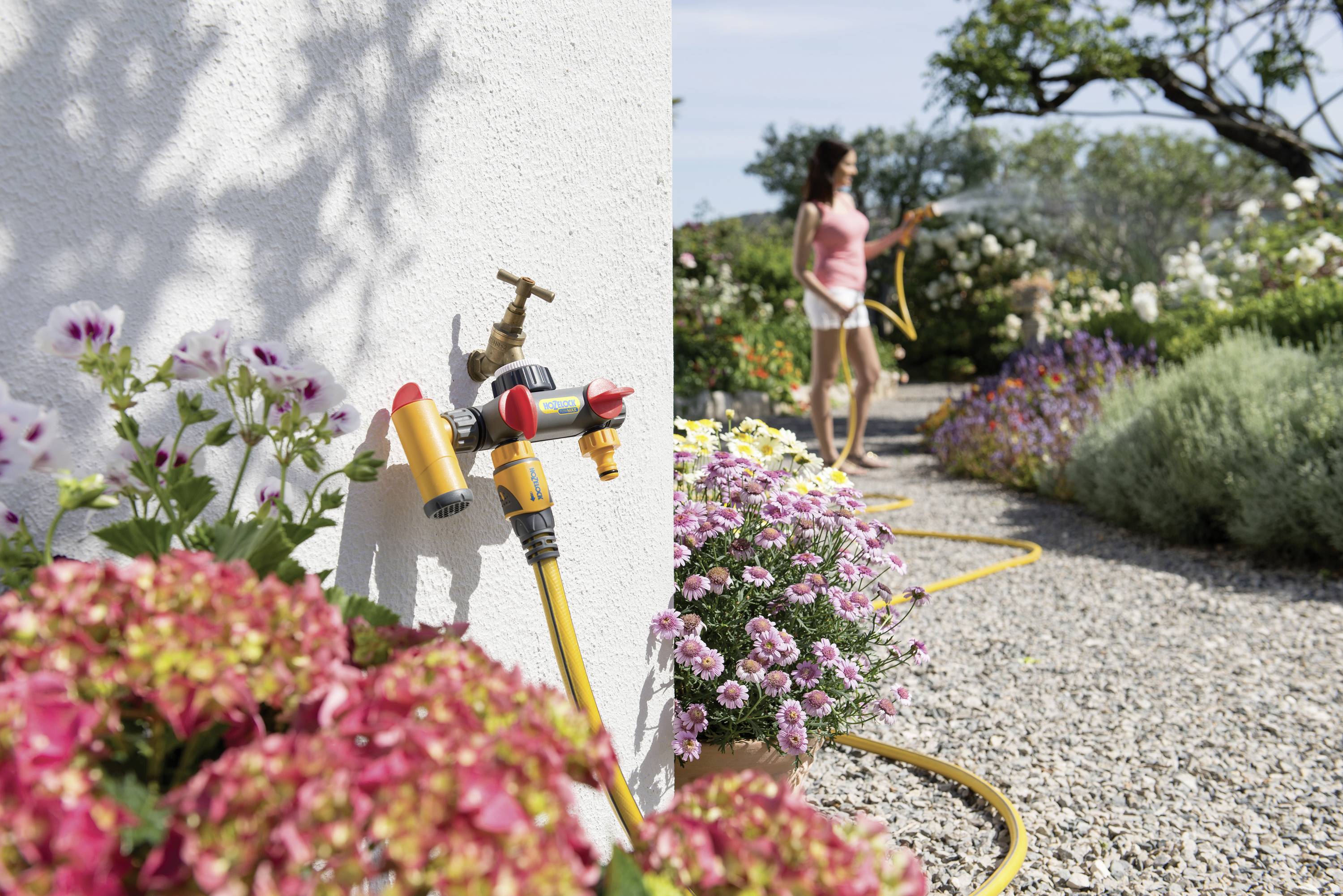 A woman waters garden plants with a hosepipe. In the foreground, a garden hose is attached to a tap.