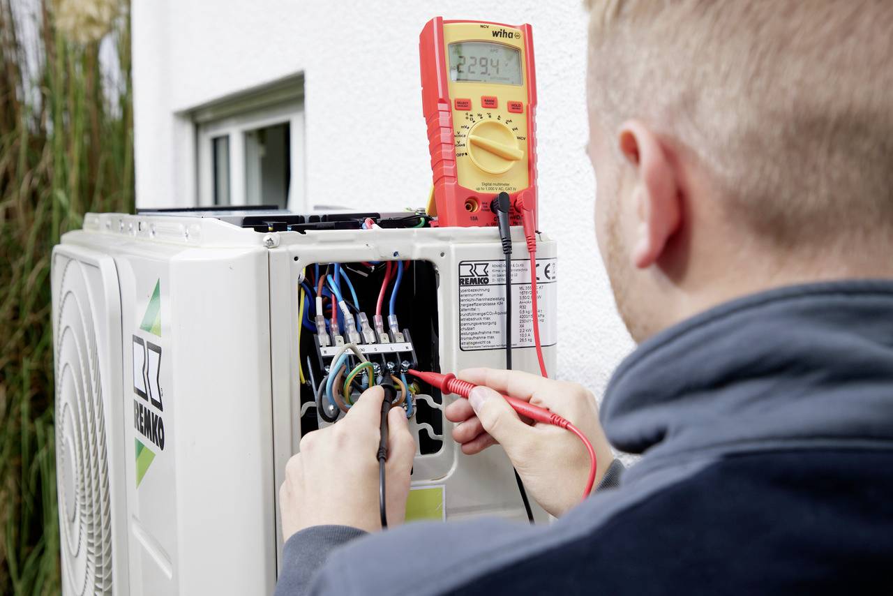 A person is measuring the voltage on an open air conditioning unit with a digital multimeter.