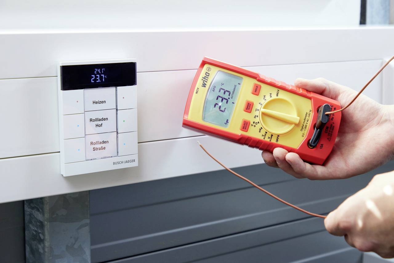 A person is measuring the voltage on a digital wall control panel using a multimetre. The display shows '22.3'.