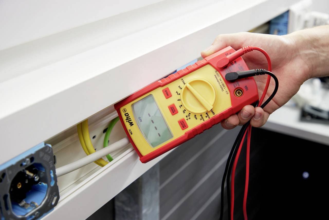 A person is holding a multimeter to test electrical wiring in a white wall panel. A blue cable is visible.