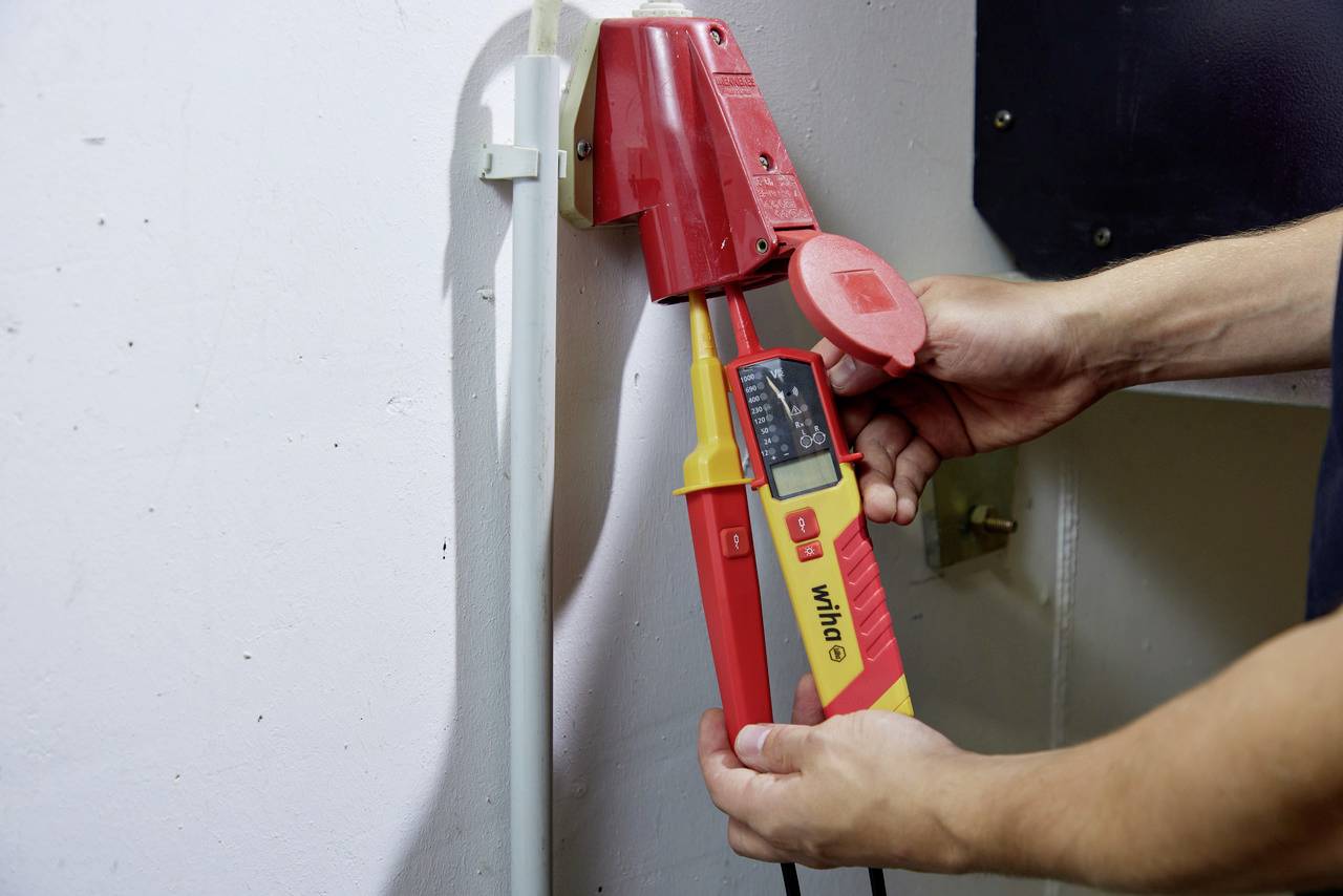 A person is testing the voltage of a red socket with a yellow multimeter, holding the probes against the connection. The measurement process is being demonstrated.