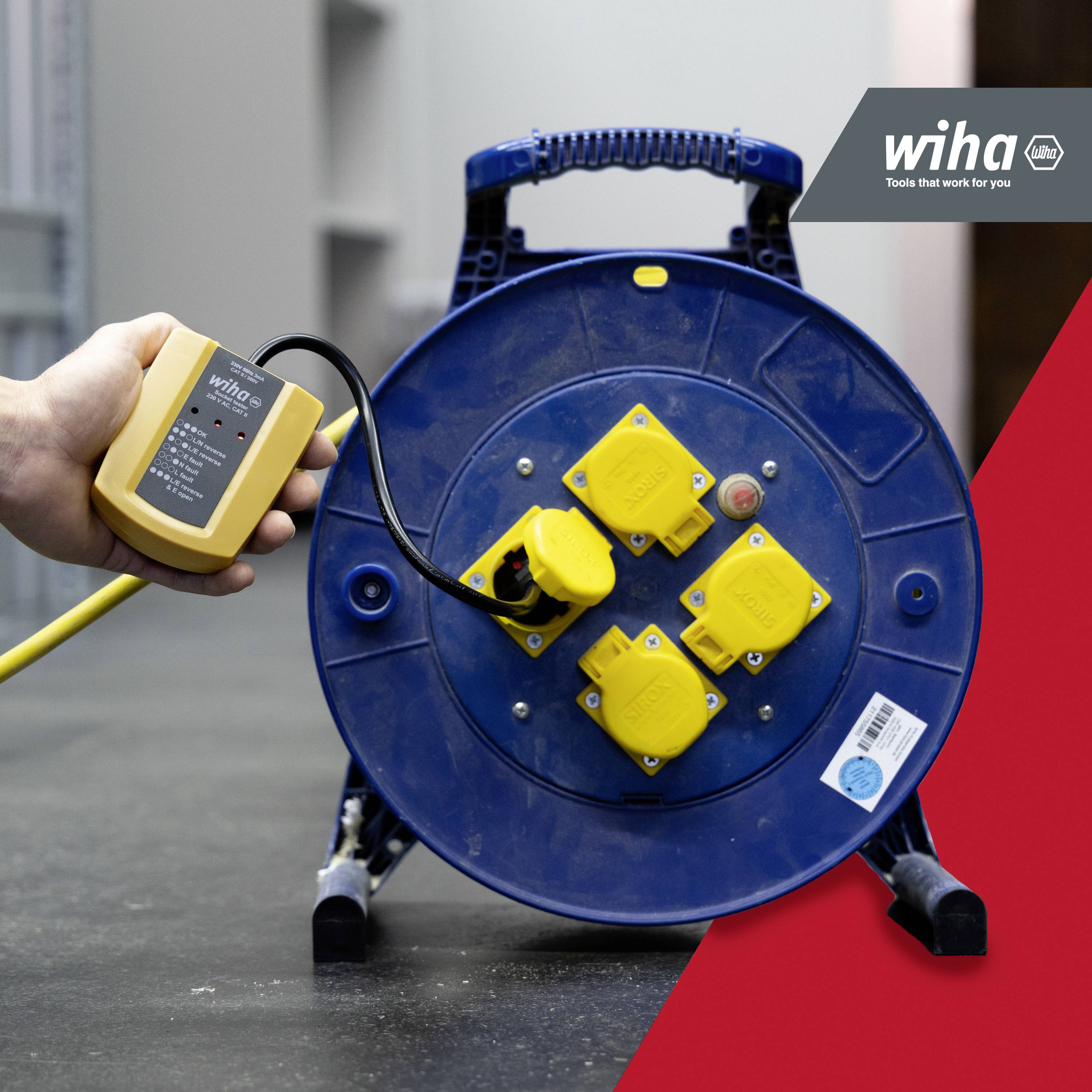 A hand holds a measuring device and tests a blue cable reel with three yellow sockets in a workshop environment.