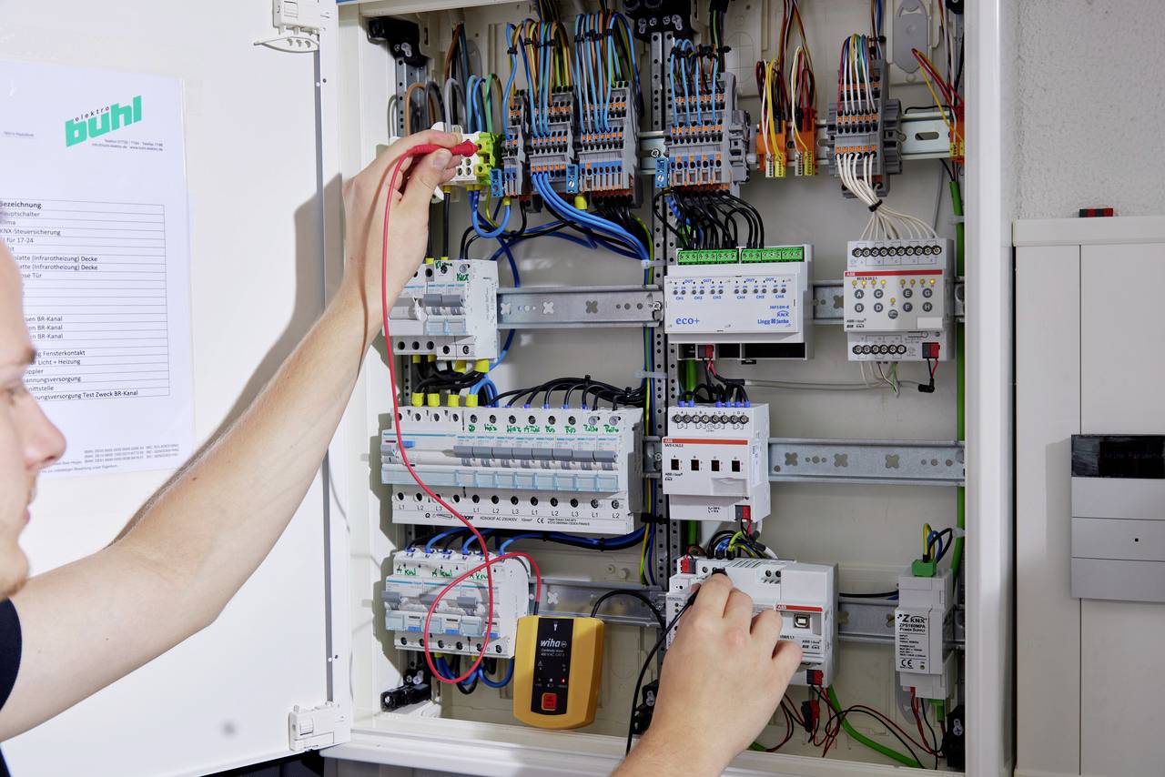 A person is working on an open electrical cabinet with numerous cables and devices, taking measurements using a multimeter.