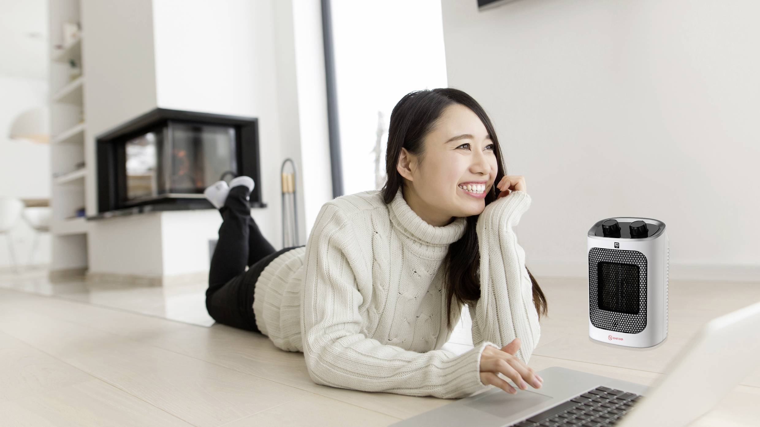 A woman in a white jumper is lying smiling on the floor and typing on a laptop. A small fan heater is next to her.