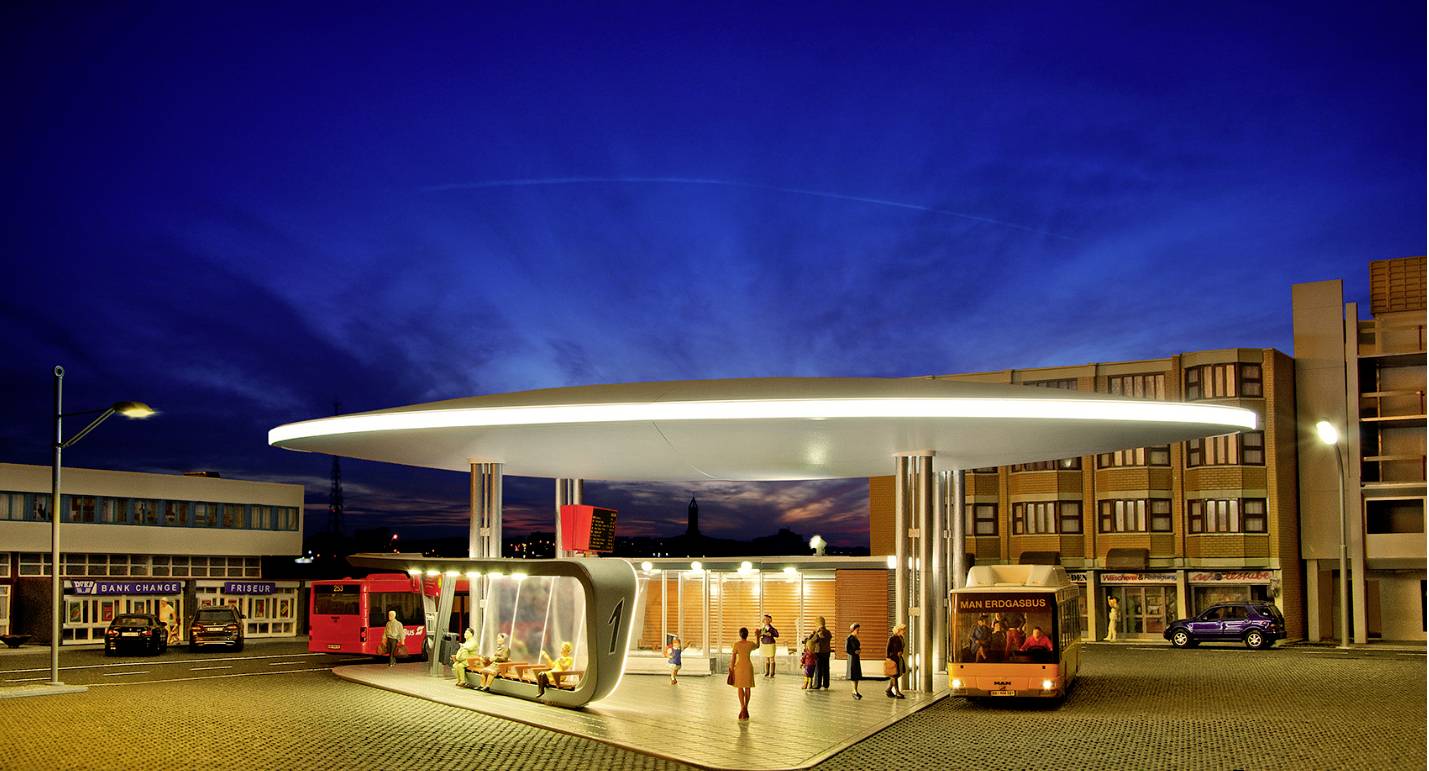 A modern, illuminated petrol station at night with several vehicles and people. A coach is parked nearby. Buildings in the background.