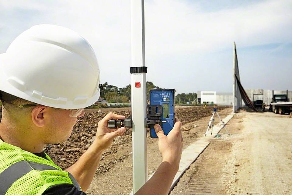 A worker wearing a hard hat and high-visibility vest is installing a measuring device on a post at a construction site. Construction work is visible in the background.