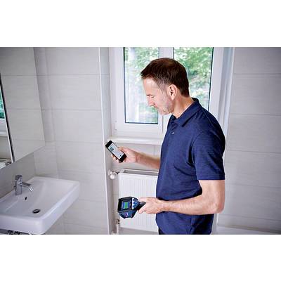 A man in a blue shirt is using a smartphone and an electronic device in a bathroom with a white tiled wall and a sink.