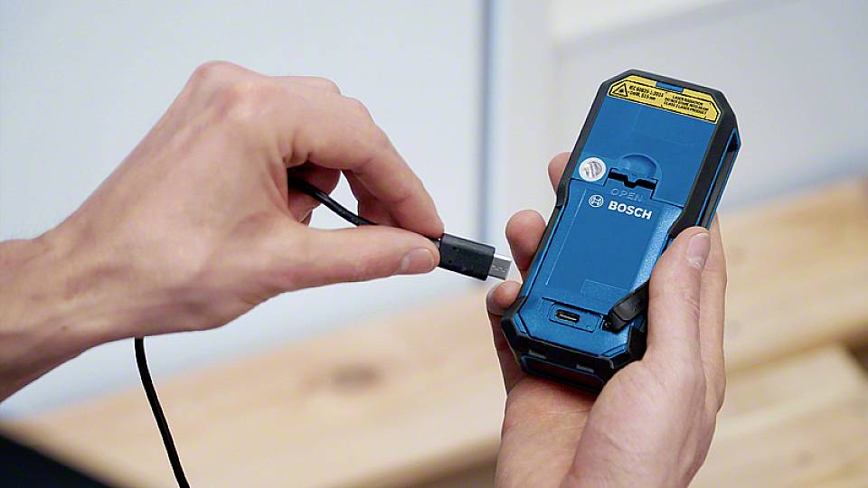 A hand is holding a blue Bosch electric tool, while the other is inserting a charging cable plug into the device.