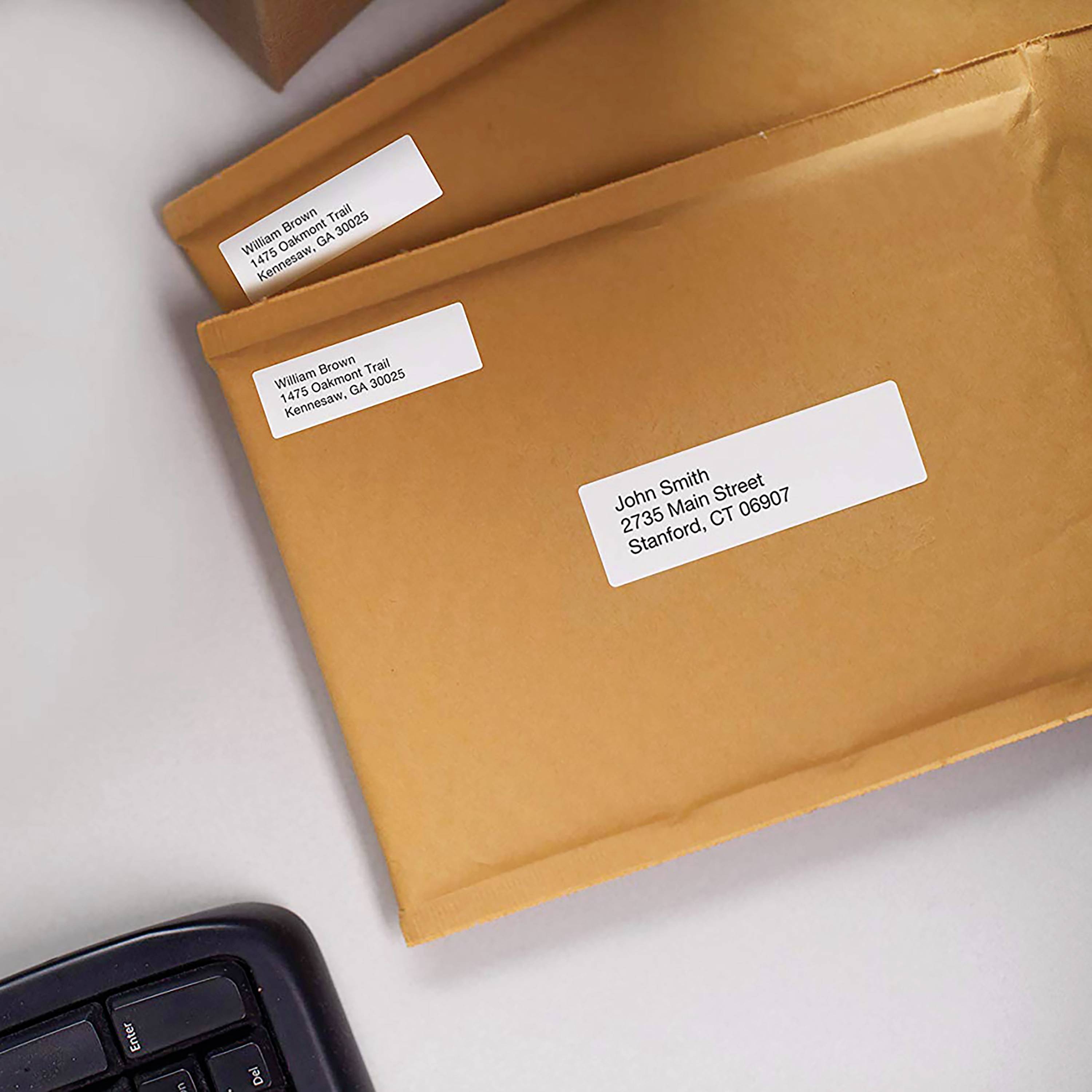 Two brown envelopes with address labels are lying on a table. The top envelope is addressed to 'John Smith, 2735 Main Street, Stamford, CT 06907'.