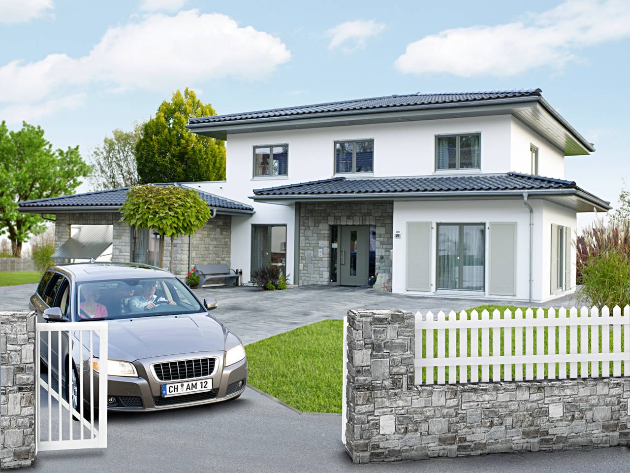 A car drives through an open gate to a modern two-storey house with a grey roof, surrounded by a garden and stone wall, under a cloudy sky.