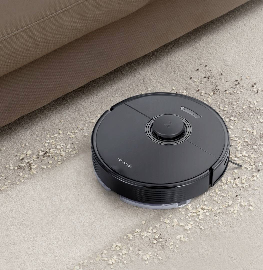 A round robotic vacuum cleaner is cleaning a beige carpet next to a brown sofa. The robot is sucking up small dirt particles.