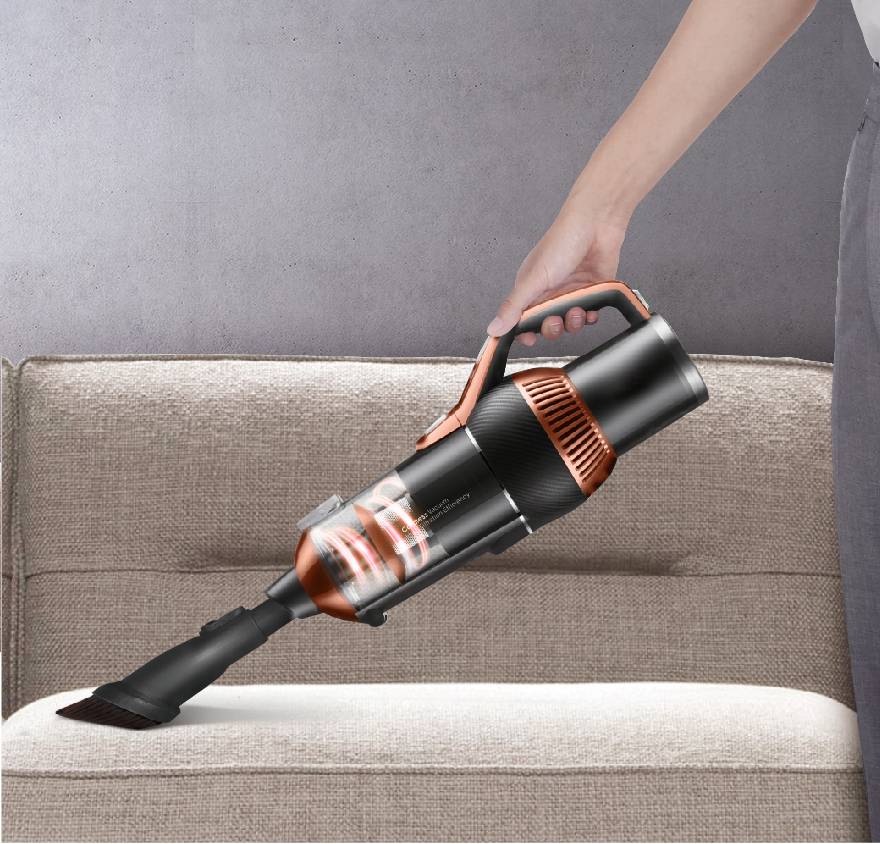 A person is cleaning a light brown sofa with a black and orange handheld vacuum cleaner. The background is a grey wall.