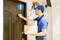 A delivery driver in a blue uniform knocks at the door and holds two parcels. A surveillance camera with a microphone is mounted above.