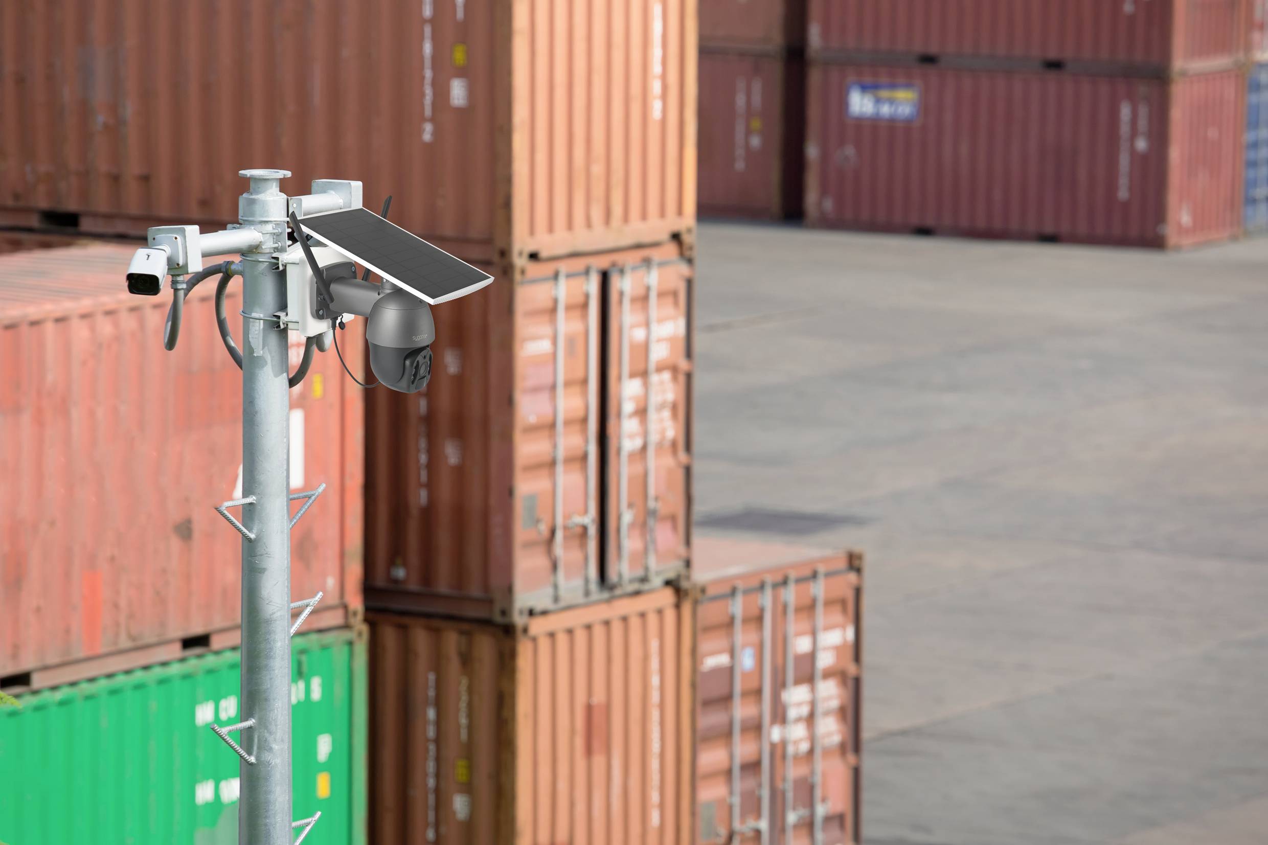 A surveillance camera with a solar module monitors a storage area with stacked freight containers in the background.