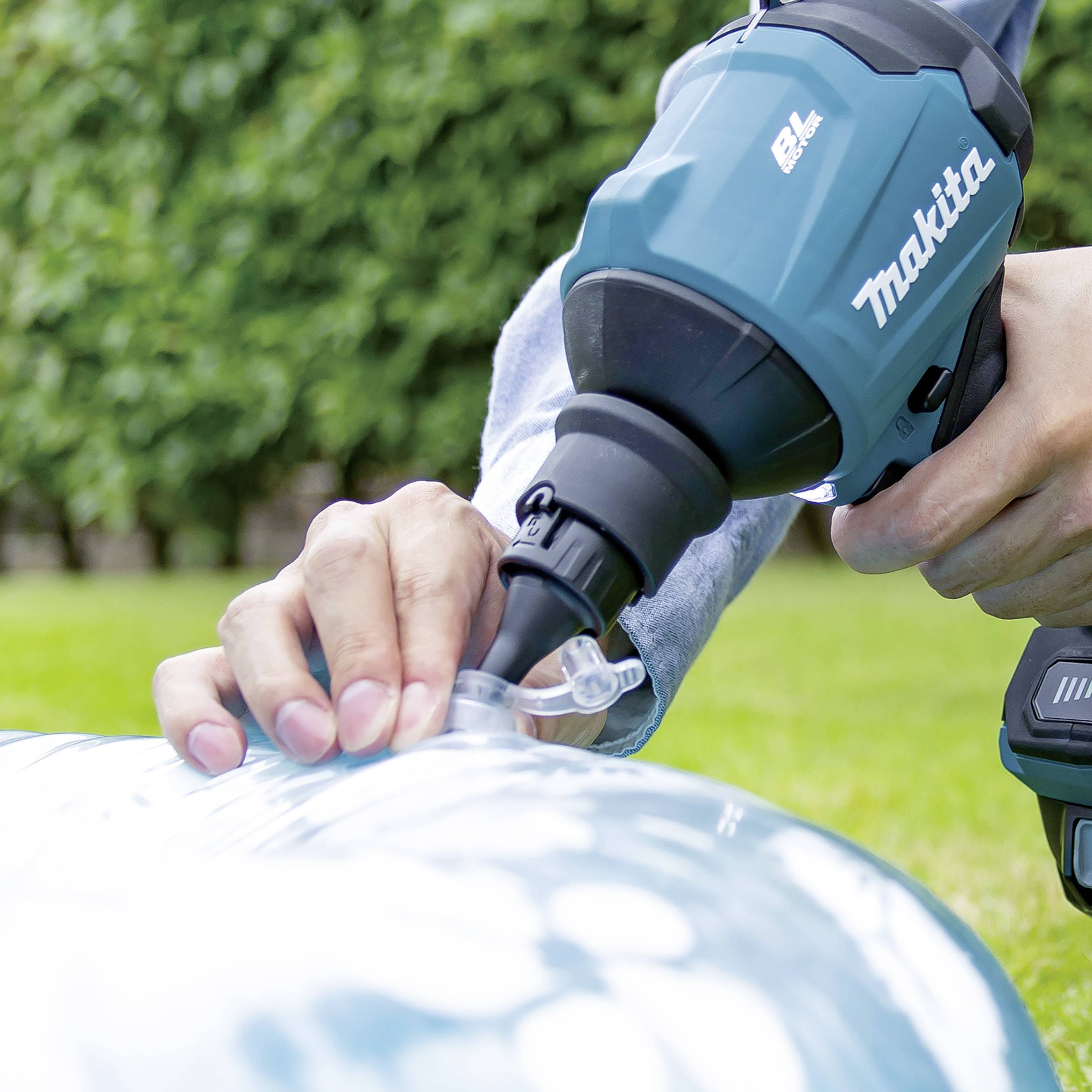 A person is using a cordless electric tool outdoors to inflate a blue balloon. Plants are visible in the background.