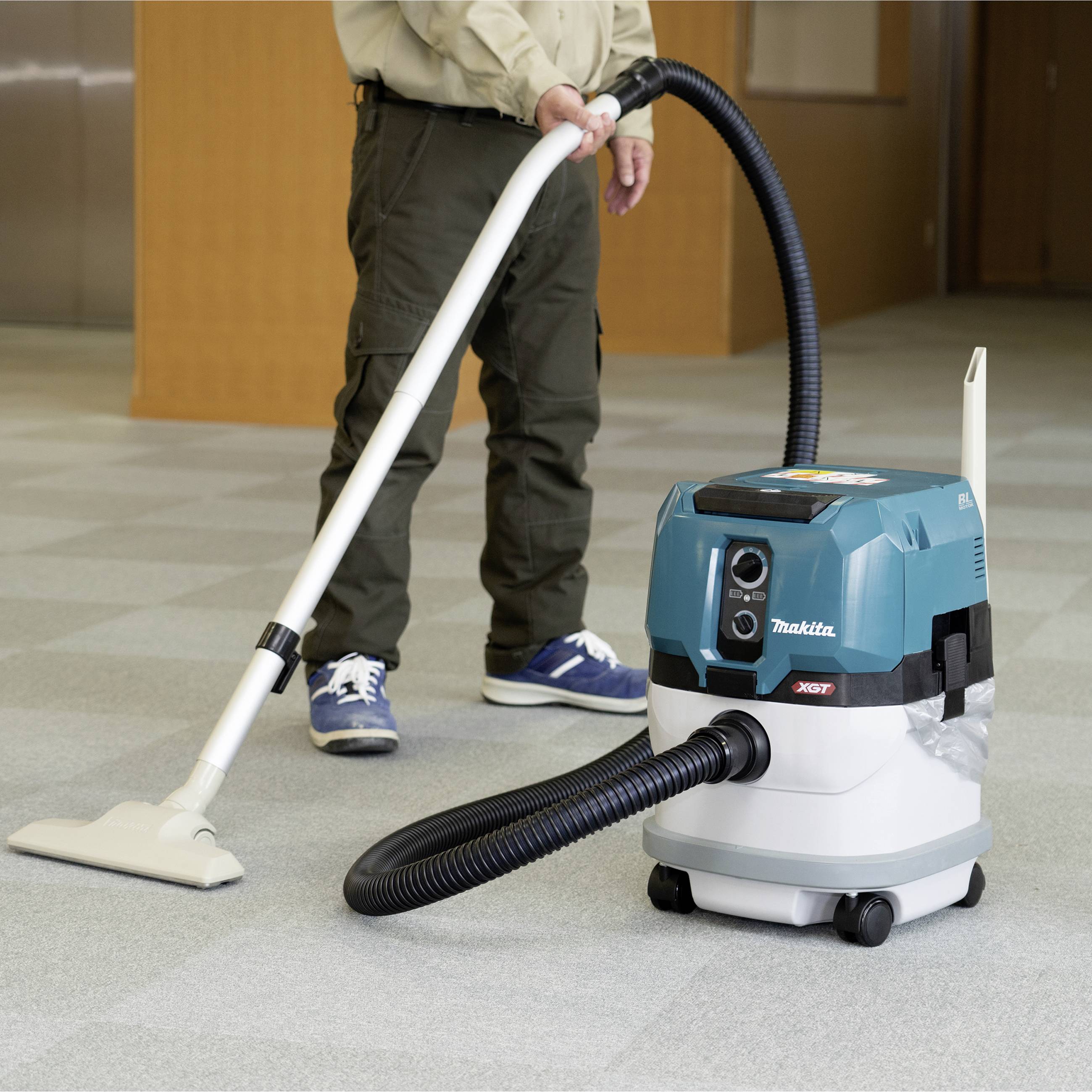 A person is vacuuming a carpeted floor with a blue and white hoover in an interior space. A wooden table is visible in the background.