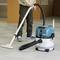 A person is vacuuming a carpeted floor with a blue and white hoover in an interior space. A wooden table is visible in the background.