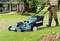 A person is mowing the lawn in front of a house with a blue Makita lawnmower. A well-maintained garden can be seen in the background.