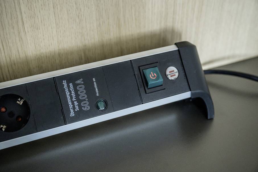 'Surge-protected power strip, 60,000A, with on/off switch and secondary button, sits on a table in front of a wooden wall.'