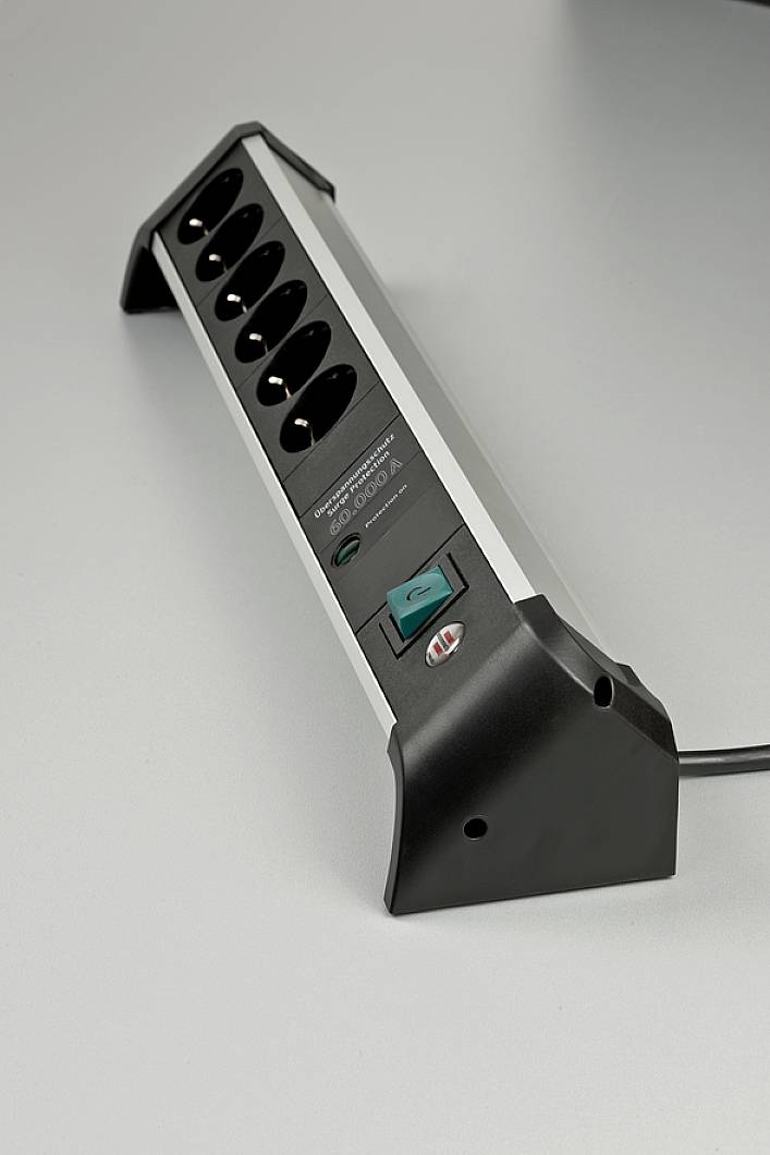 A black multi-socket extension with six plug sockets and an illuminated on/off switch is lying on a grey surface.
