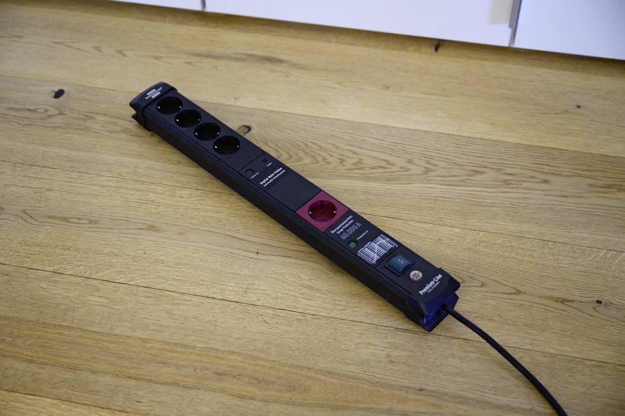 A black multi-socket extension lead with five sockets and a red-marked master switch lies on a wooden floor beneath white cupboards.