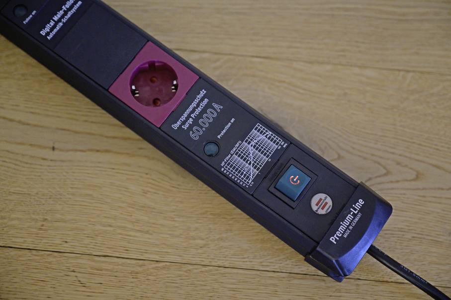 A black premium-line power strip with a red socket and a green switch is lying on a wooden floor.