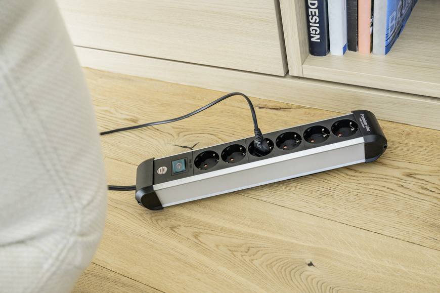 A black power strip with six socket outlets and a green power switch lies on a wooden floor in front of a bookshelf. One device is plugged in.