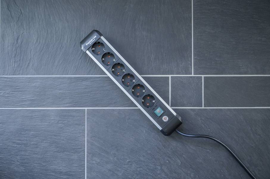 A black six-socket extension lead is positioned at an angle on a grey tiled floor. One cable is plugged in.