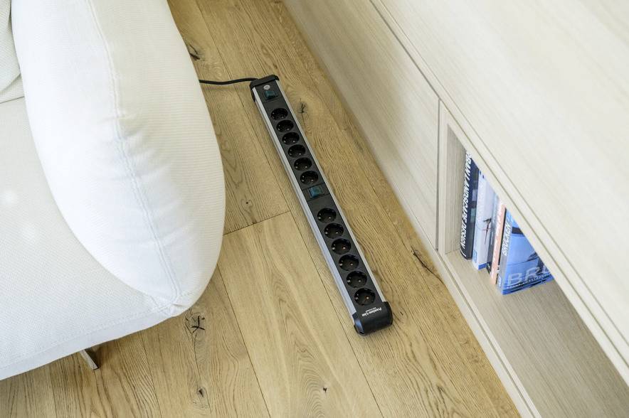 A multi-socket extension lead is lying on the wooden floor next to a sofa and a bookshelf.
