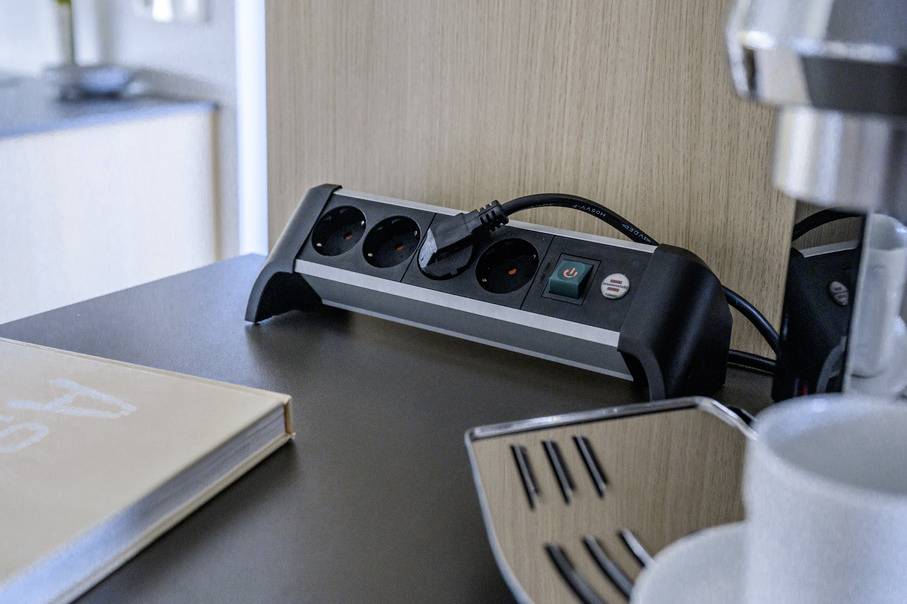 A multi-socket extension lead with switches and sockets sitting on a table next to a book and a coffee machine in a room.