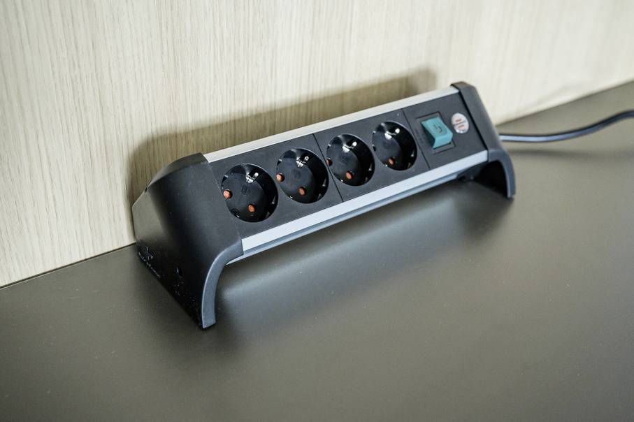A four-socket power strip with a rocker switch is lying on a table in front of a wooden wall.