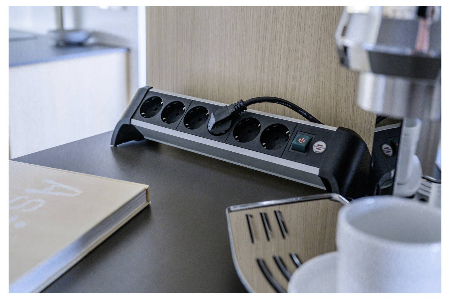 Power strip with six outlets and a plugged-in cord on a desk beside a coffee machine and mug, suggesting a workspace or break area.
