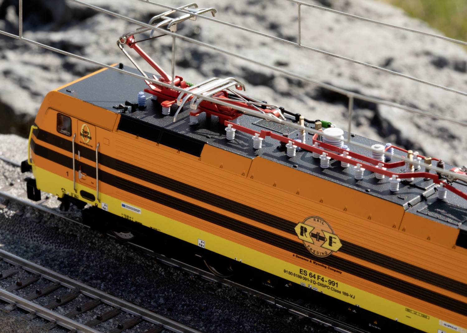 Toy model of a yellow and black electric locomotive with a detailed view of the overhead line, positioned on tracks against a rocky background.