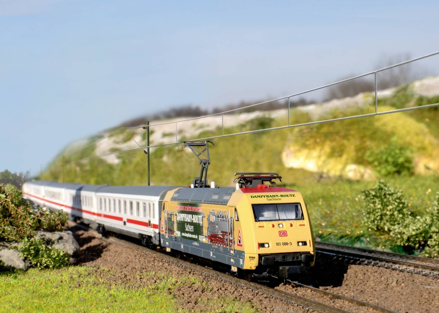 A model train travels along a railway track through a scenic landscape with green hills and a blue sky in the background.