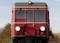 Red train from the front against a clear sky. Vehicle stationary, front view. Background shows autumnal trees.