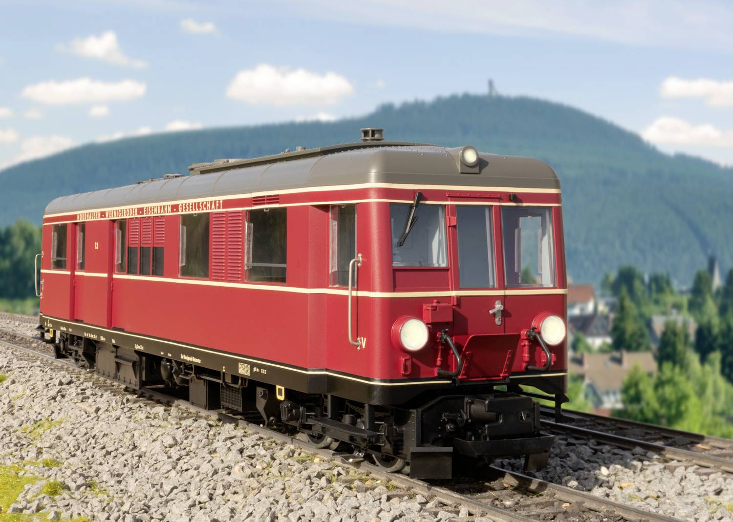 A red, historic railcar travels along tracks through a hilly landscape with a wooded hill in the background.