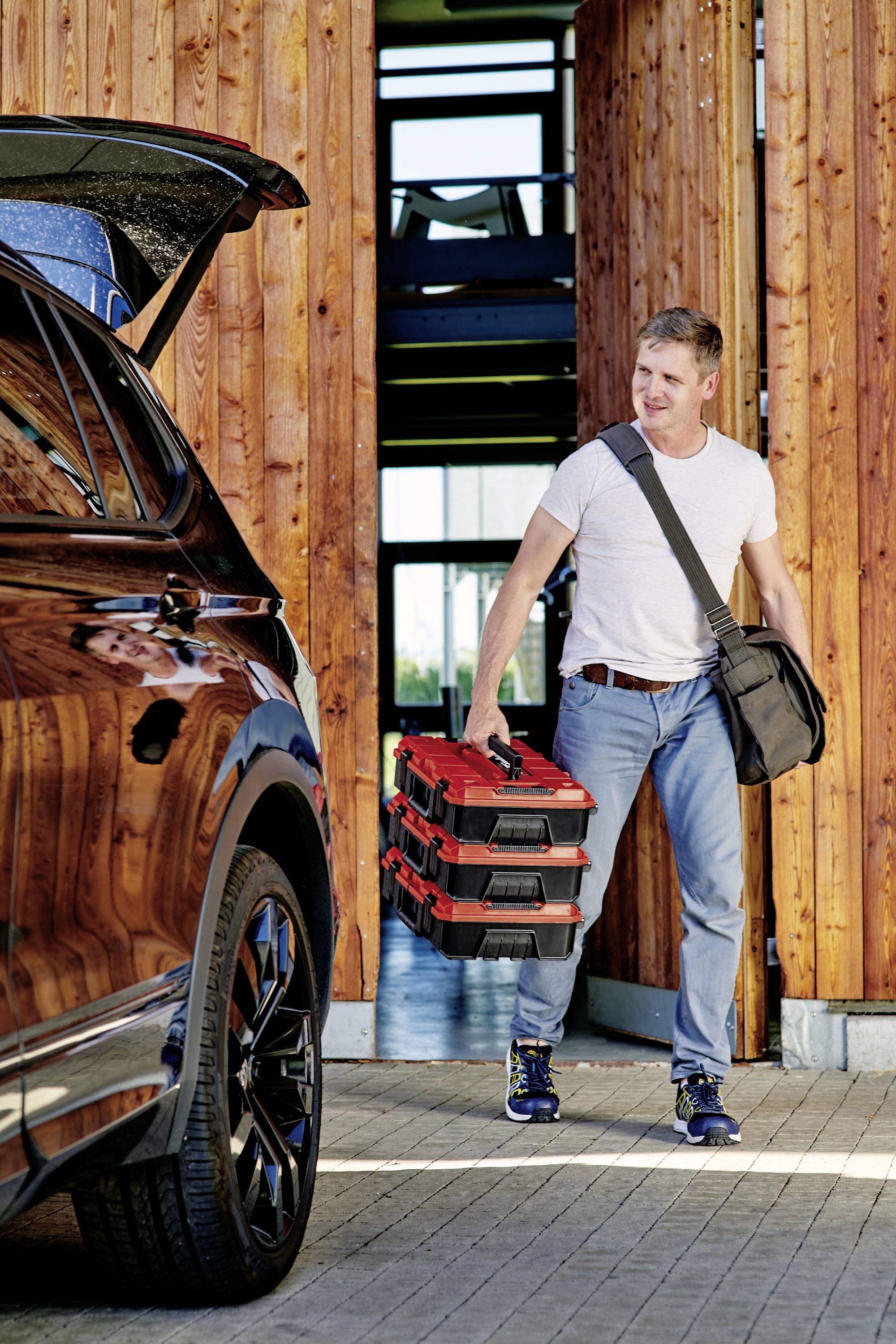 A person wearing jeans and a white T-shirt is carrying two red toolboxes and walking towards a car. The surroundings are modern with wooden panelling.