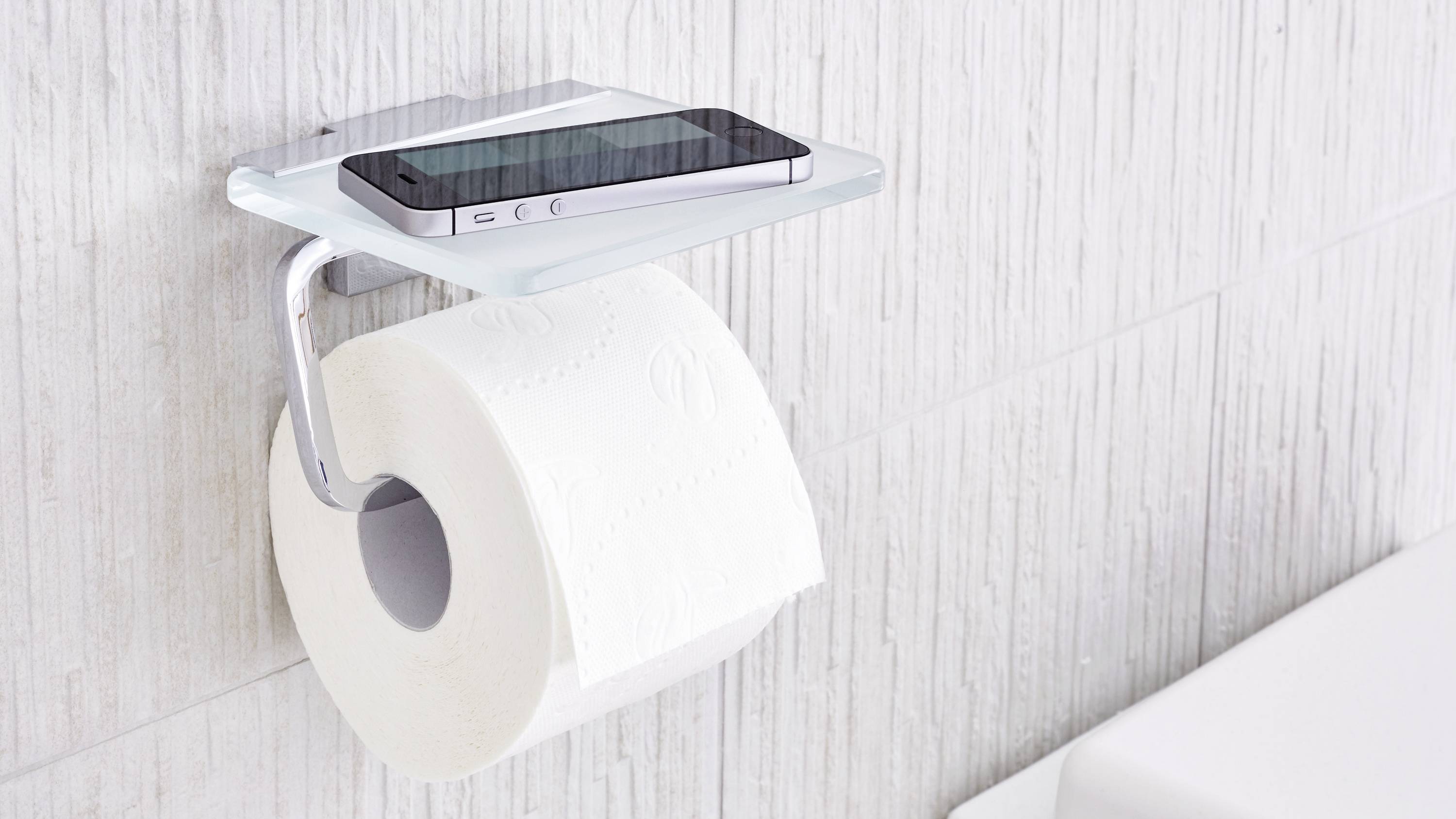 Toilet roll holder with an integrated shelf for a smartphone mounted on a white tiled wall.