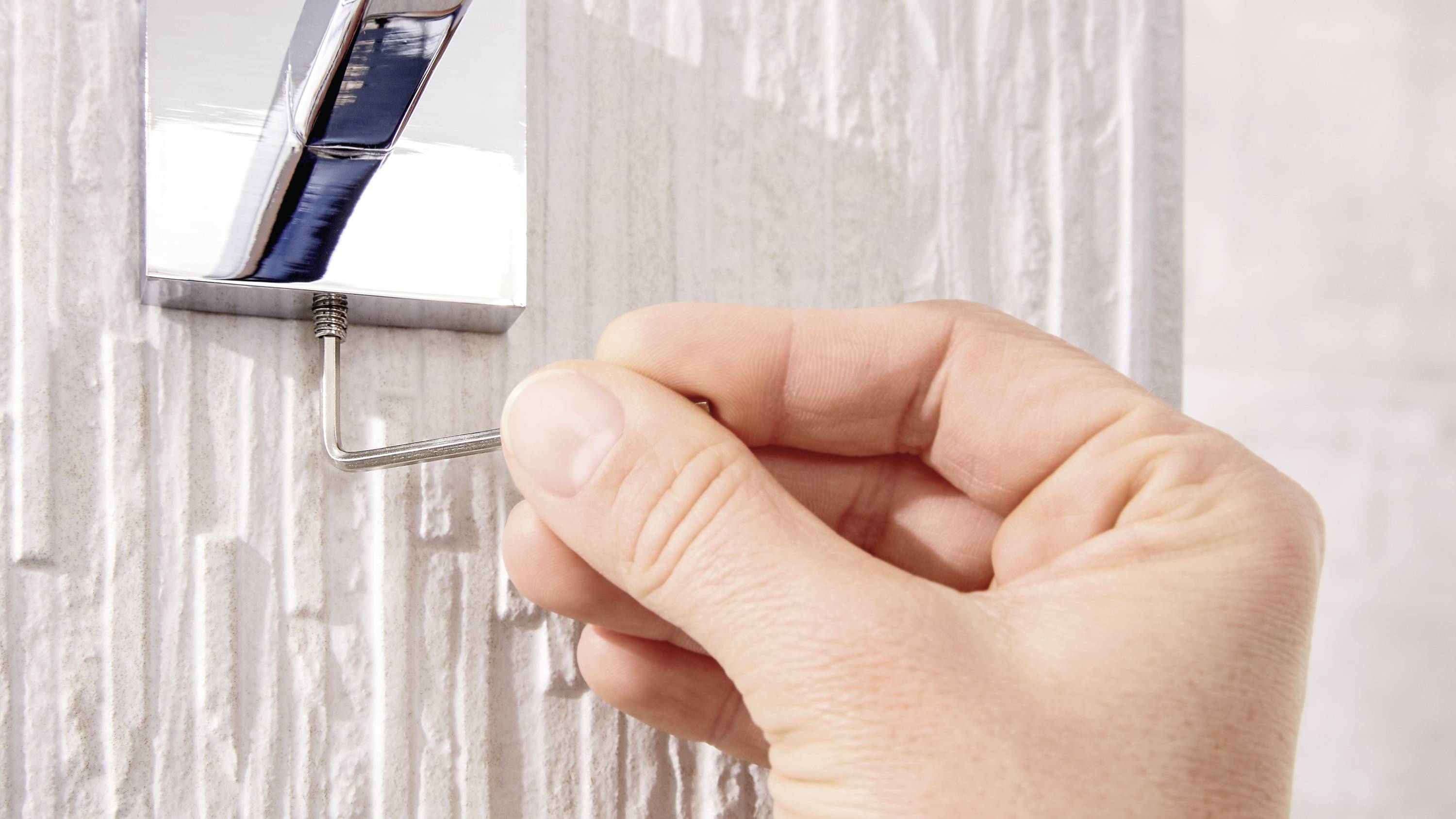 A hand is tightening a screw with an Allen key to secure a fitting to a wall.