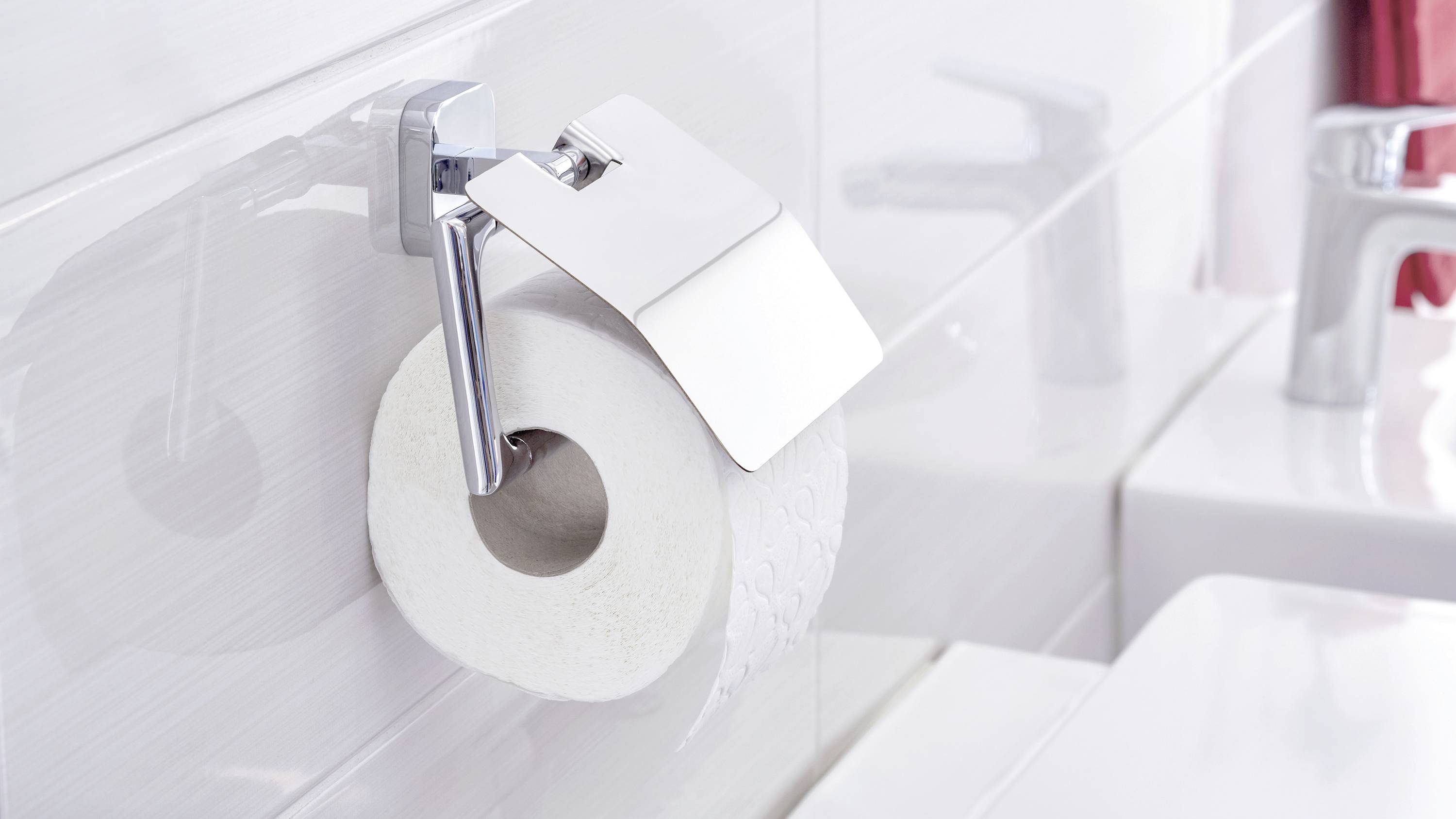 A toilet with a white toilet roll holder, on which a toilet roll hangs; next to it a washbasin with a tap.